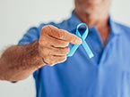 Torso of older male nurse in blue scrubs holding blue ribbon for prostate cancer awareness in his right hand