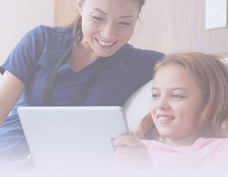 Female nurse in blue scrubs sitting on bed with girl patient holding a tablet representing pediatric nursing CEUs