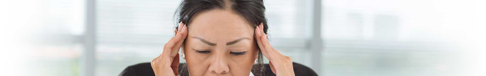 Asian woman in light pink blouse sitting at desk holding her hands to her temples trying to manage her pain 