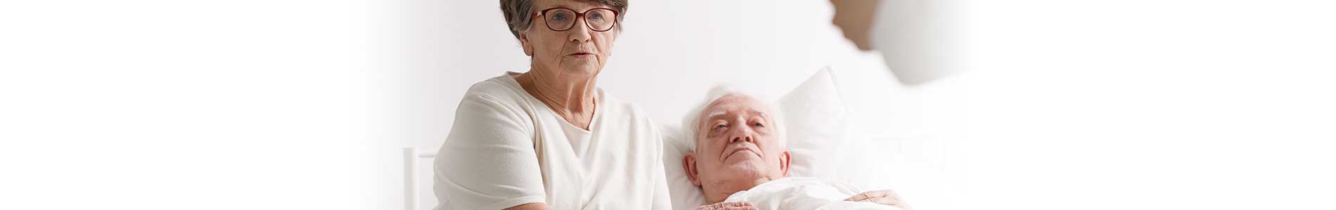 Elderly woman is caringly holding older male adult's hand who is laying in bed for end-of-life while listening to Nurse