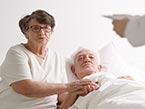 Elderly woman is caringly holding older male adult's hand who is laying in bed for end-of-life while listening to Nurse