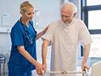 Female nurse in blue scrubs preventing elderly male patient in gown with walker from falling