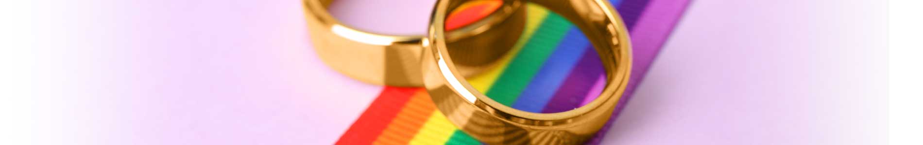 Two golden wedding rings laying on rainbow ribbon on pink background for LGBTQ awareness