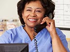 Black older nurse in blue scrubs behind computer screen holding a phone receiver for a telephone triage call