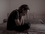Young man in a mental health crisis sitting cross-legged on bed holding his hands in front of his face