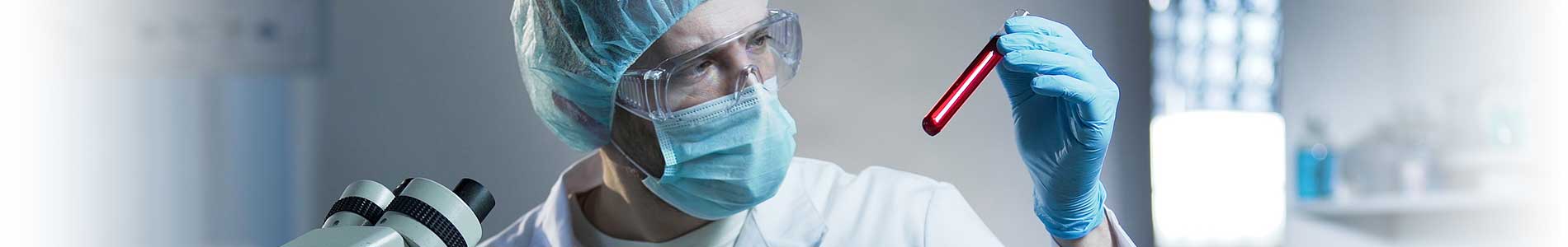 Medical professional with lab coat, face mask, gloves and hair net, holding and looking at blood testing tube for pathogens