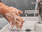 Man in white infection control protection suit wearing goggles holding Ipad in one hand and raised hands towards viewer