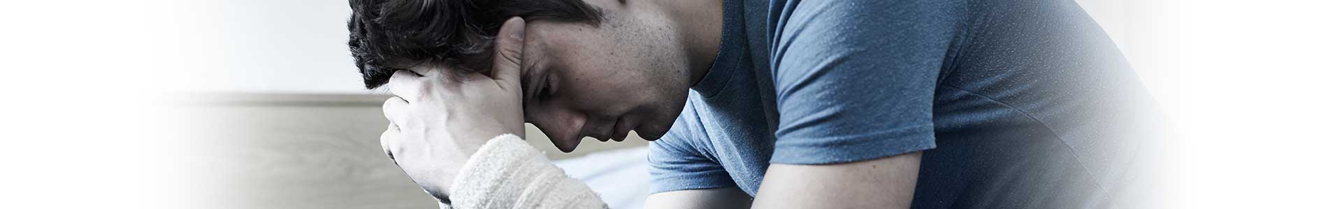 Stressed and depressed man in blue jeans and t-shirt sitting on bed with face in his hands and bandage on wrists