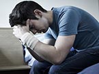 Stressed and depressed man in blue jeans and t-shirt sitting on bed with face in his hands and bandage on wrists