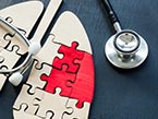Plain-wooden puzzle pieces in shape of lungs with 2 red puzzle pieces representing cancer below a stethoscope