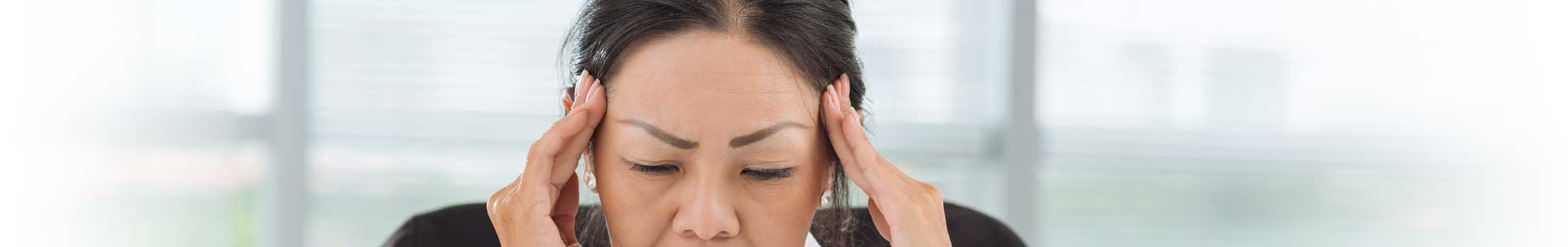 Asian woman in light pink blouse sitting at desk holding her hands to her temples trying to manage her pain