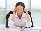 Asian woman in light pink blouse sitting at desk holding her hands to her temples trying to manage her pain