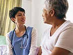 Nurse in blue scrubs and stethoscope talking to elderly female patient in t-shirt sitting on bed about health