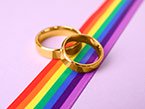 Two golden wedding rings laying on rainbow ribbon on pink background for LGBTQ awareness