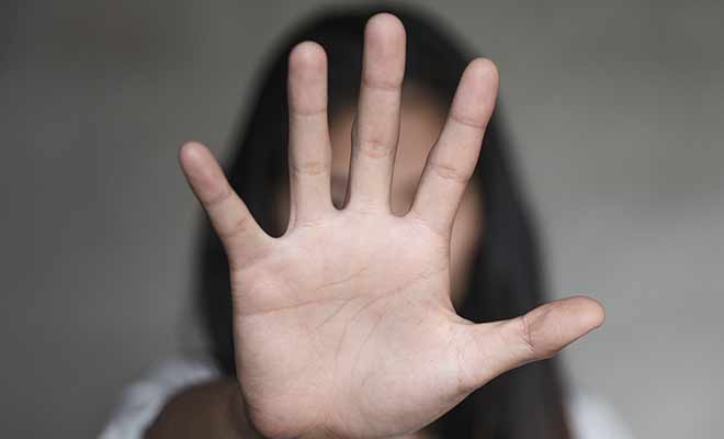 Woman with long dark hair in white shirt with raised hand covering her face to prevent herself from sexual harassment